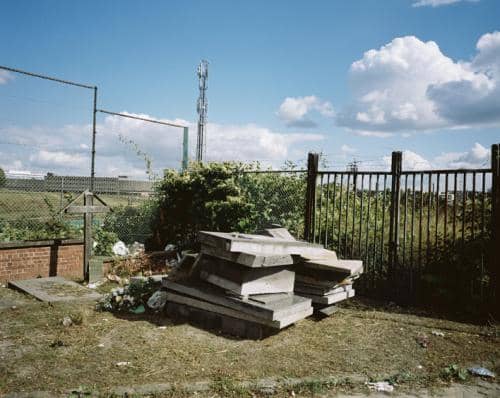 Un dépôt dans le fond d'un cimetière à Bruxelles, des pierres tombales entassées, une vieille croix en bois le tout sur fond de grillage et de barbelés, photographie argentique moyen format Plauble W67