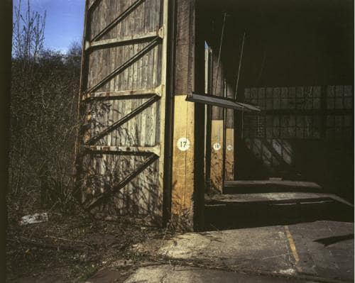 un hangar dépôt de trains abandonné avec des portes en bois, de la peinture défraichie et des railles rouillées, le tout donnant sur la nature sauvage, photographie argentique moyen format Plauble W67
