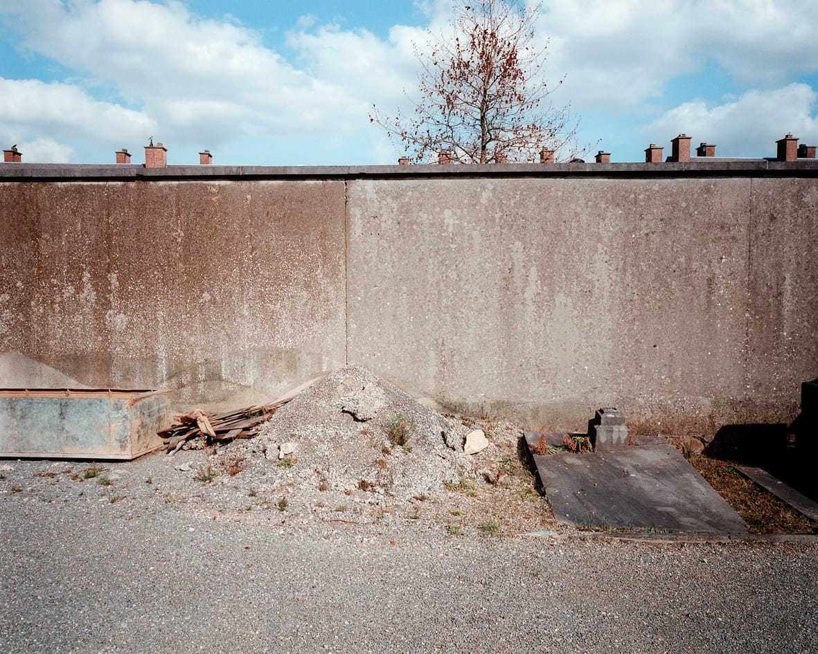 Un cimetière abandonné avec de vieilles pierres tombales et un tas de sable donnant sur une cité dortoir moderne derrière les murs, photographie argentique moyen format Plauble W67